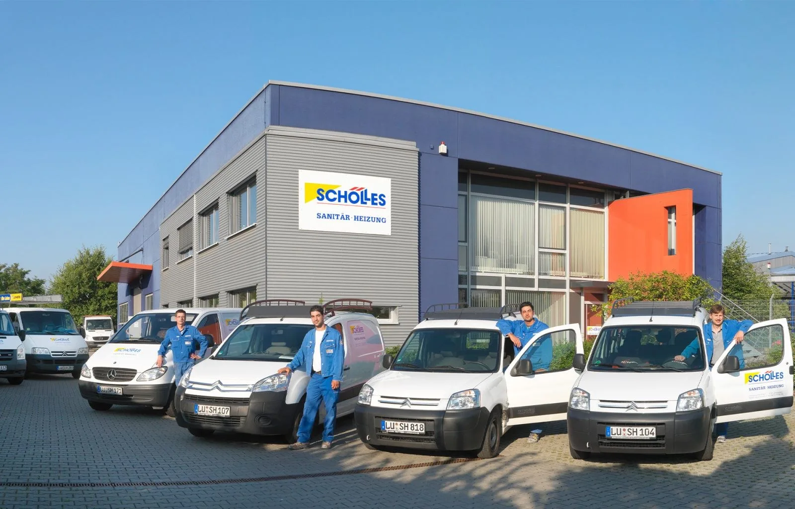 Vier Monteure in blauen Schölles-Overalls neben weißen Firmentransportern vor dem Firmengebäude mit dem Schild Schölles Sanitär Heizung