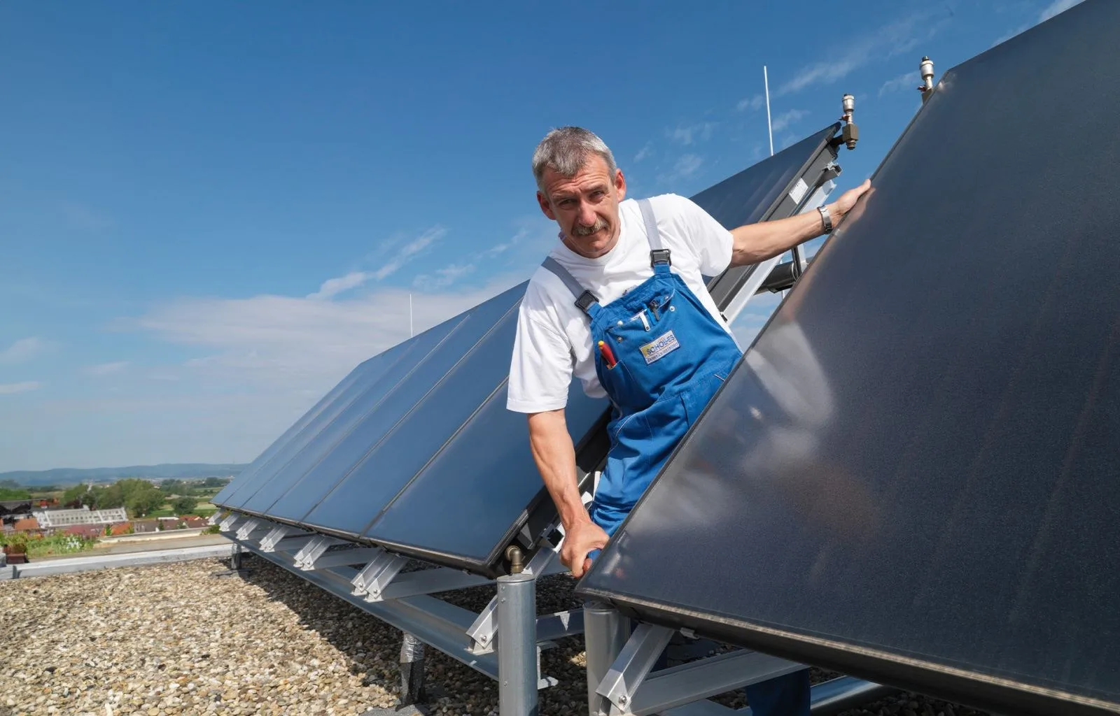 Monteur mit Schölles-Aufnäher an der Latzhose lehnt an Solarkollektoren auf einem Flachdach
